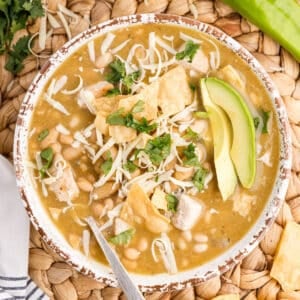 Bowl of green chicken chili garnished with cilantro, avocado slices, melted cheese, on a woven mat with green chiles and lime halves.