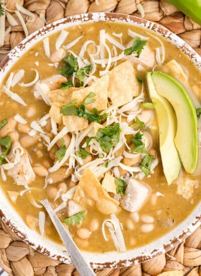 Bowl of green chicken chili garnished with cilantro, avocado slices, melted cheese, on a woven mat with green chiles and lime halves.