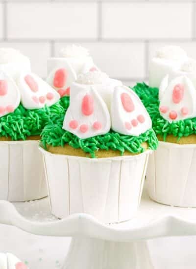 View of Bunny Butt Cupcakes on a platter showing the underside of the bunnies feet, with other cupcakes in the background and text overlay.