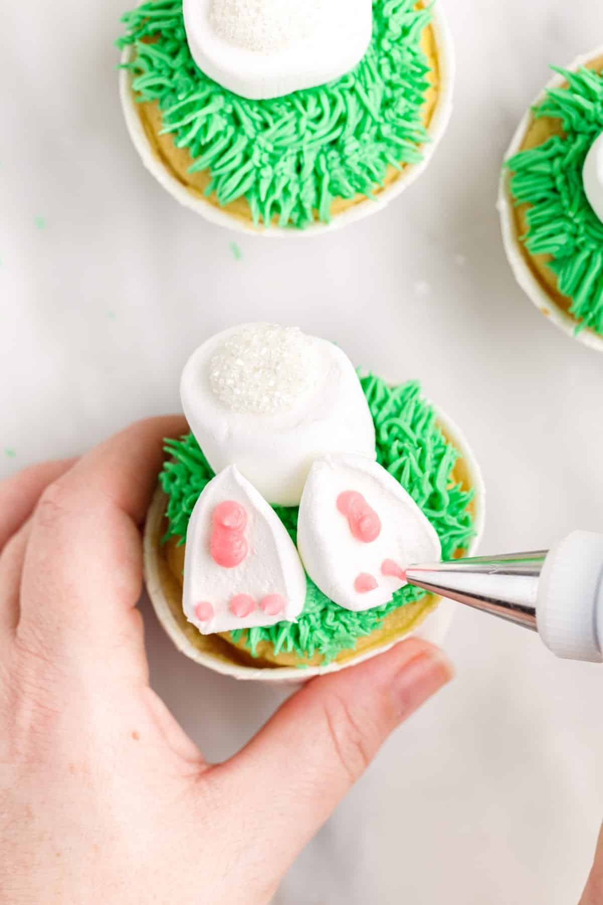 Pipping the pink pads of the bunny feet onto the marshmallows, with other cupcakes in the background.