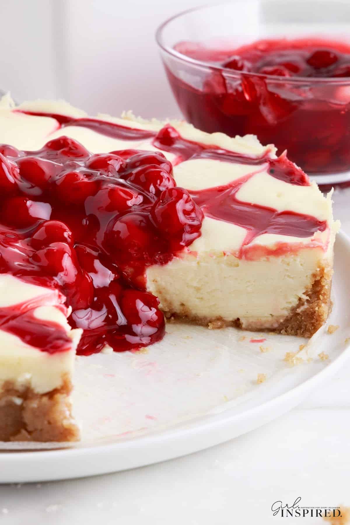 Side view of the Cherry Cheesecake showing the graham cracker crust, cheesecake filling and cherries oozing over the side, with a slice taken out so you can see the insides of the cake.