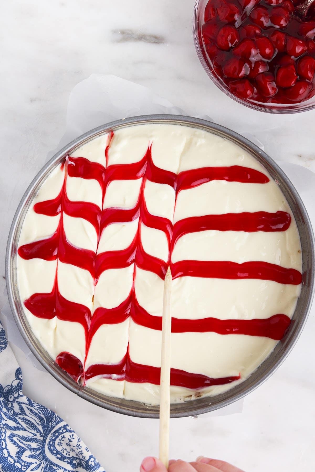 Using a stick, making the pattern in the cherry sauce on top of the cheesecake layer. with cherries in the background.