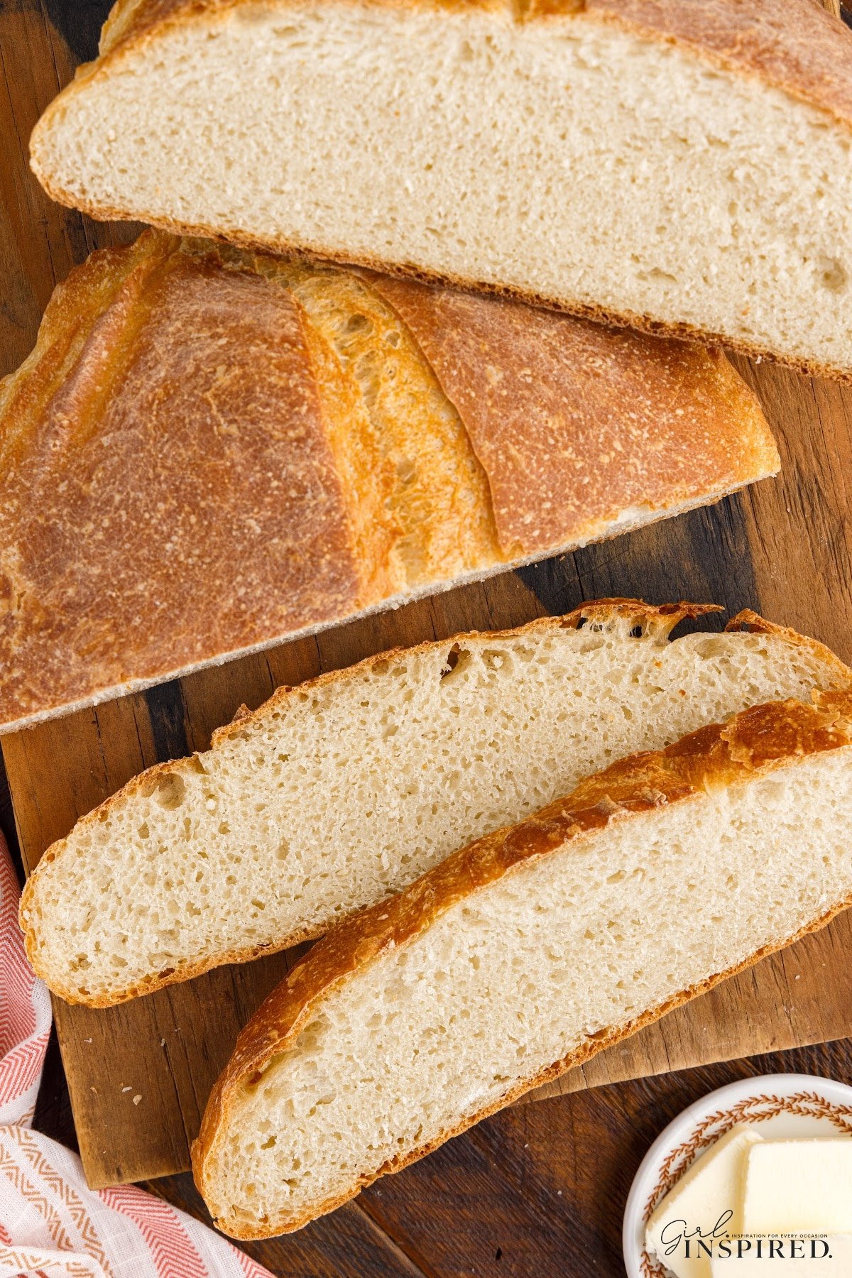 Slices off a loaf of Dutch Oven Bread on a wooden cutting board, with other ingredients in the background.