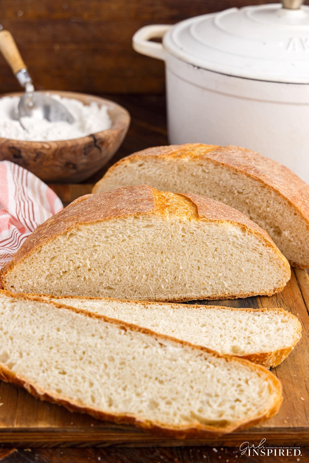 Slices off a loaf of Dutch Oven Bread on a wooden cutting board, with other ingredients in the background.