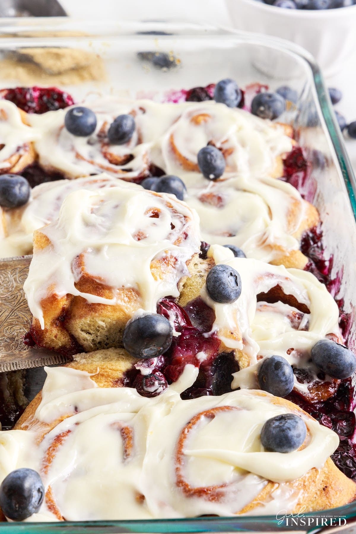 Close up of Blueberry Cinnamon Rolls in a baking dish with one on a spatula, showing the blueberries and frosting.