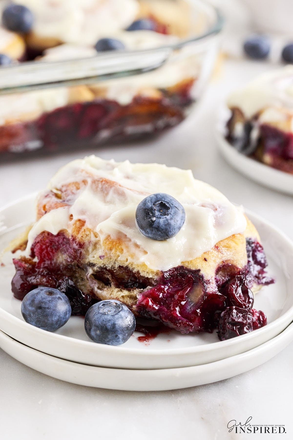 One piece of Blueberry Cinnamon Rolls on a plate, with a dish filled with rolls in the background.