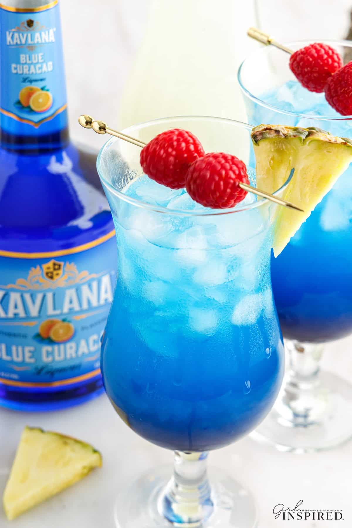 Close up of two tall glasses with fruit tingle cocktail and garnished with pineapple wedges and skewered raspberries, bottle of Blue Curacao, pineapple wedge garnish on a marble countertop.