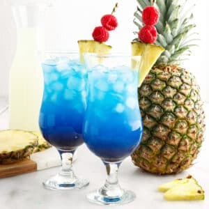Elevated shot of two tall glasses filled with fruit tingle cocktail and garnished with pineapple wedges and skewered raspberries, whole pineapple standing in an upright position, a bottle of Blue Curacao, pineapple wedge on a marble countertop.