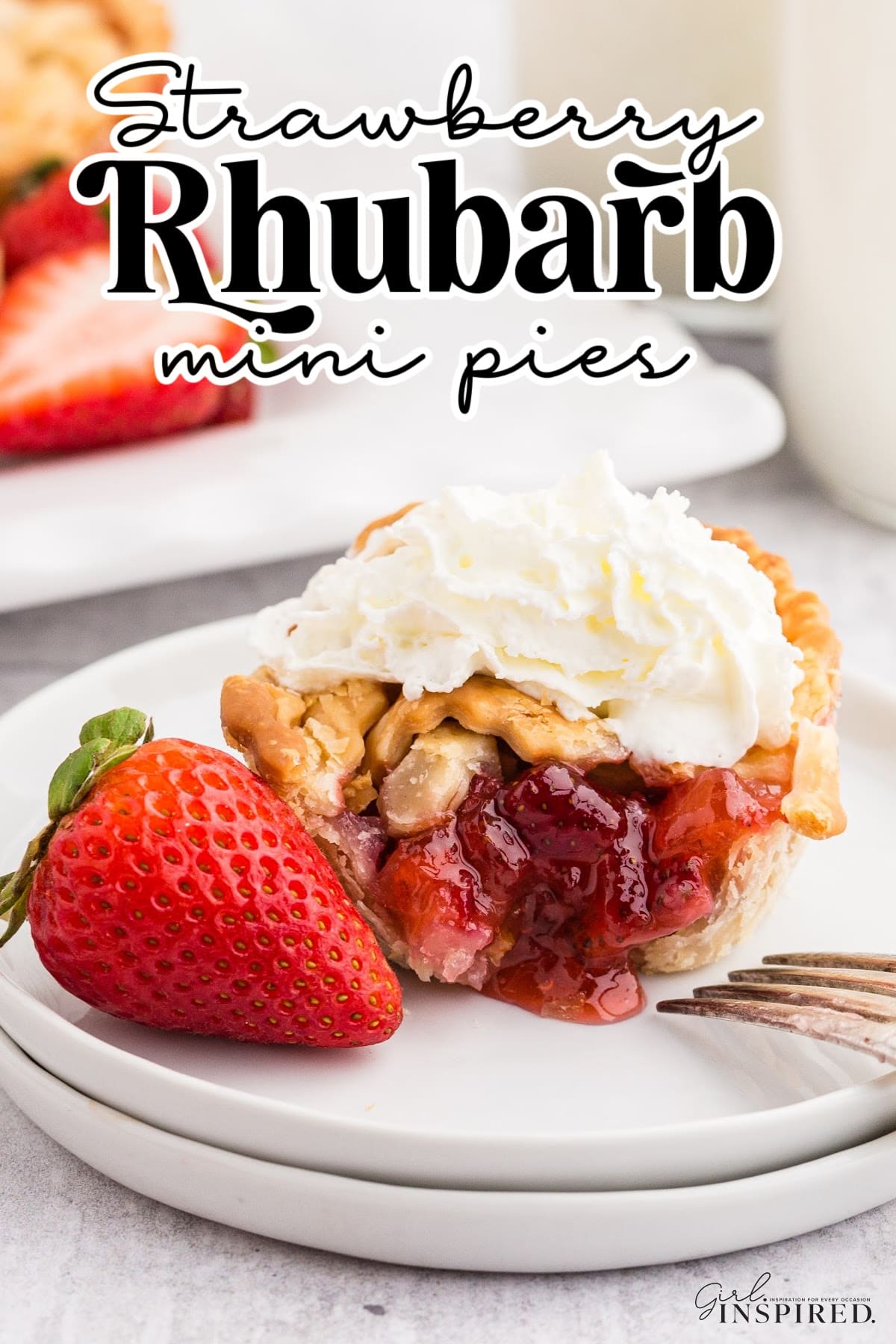 Strawberry Rhubarb mini pie on a plate, cut open to show the strawberry inside, topped with whipped cream and a whole strawberry on the plate, with other mini pies in the background., with text overlay.