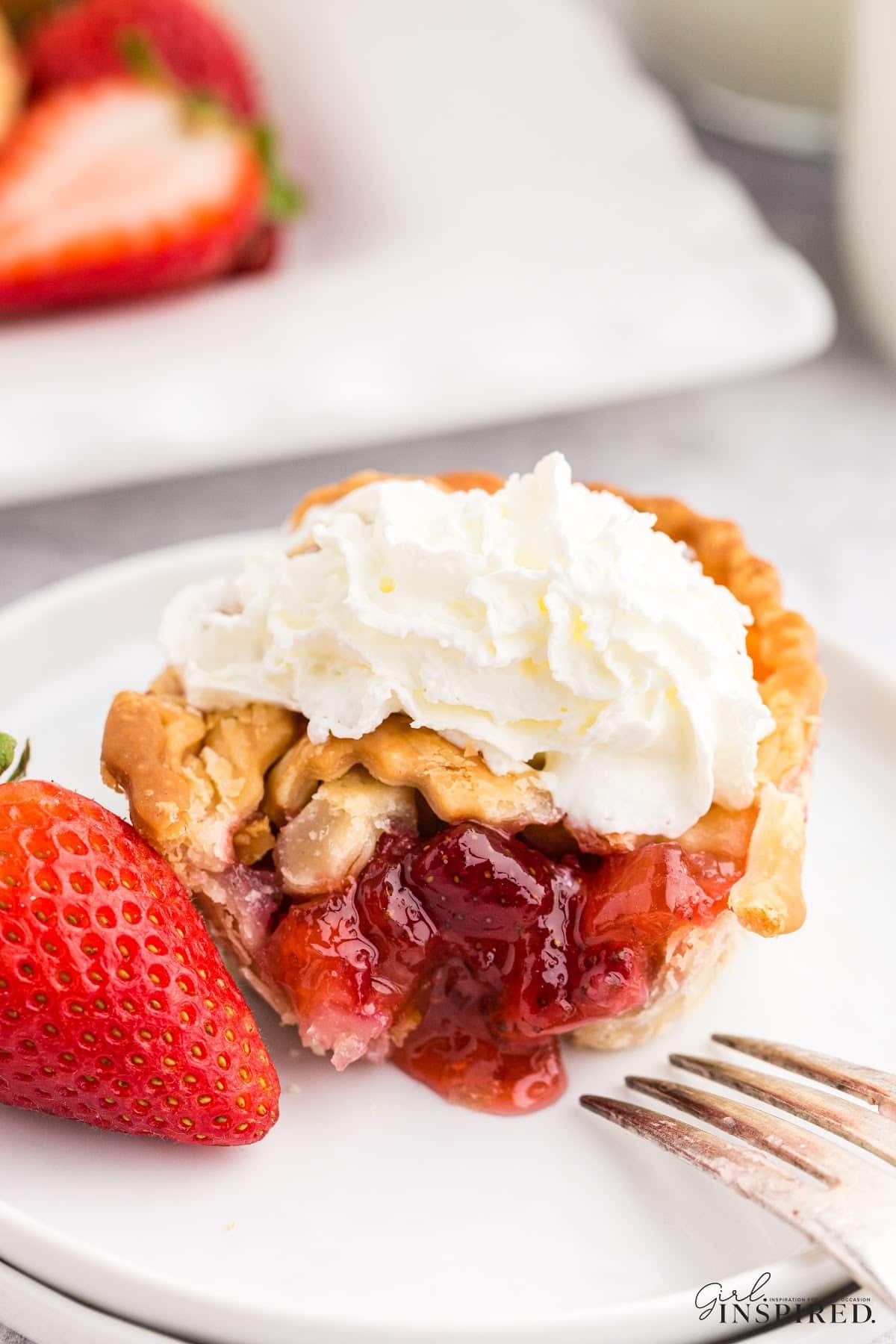 Strawberry Rhubarb mini pie on a plate, cut open to show the strawberry inside, topped with whipped cream and a whole strawberry on the plate, with other mini pies in the background., with text overlay.