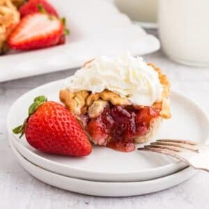 Strawberry Rhubarb mini pie on a plate, cut open to show the strawberry inside, topped with whipped cream and a whole strawberry on the plate, with other mini pies in the background., with text overlay.