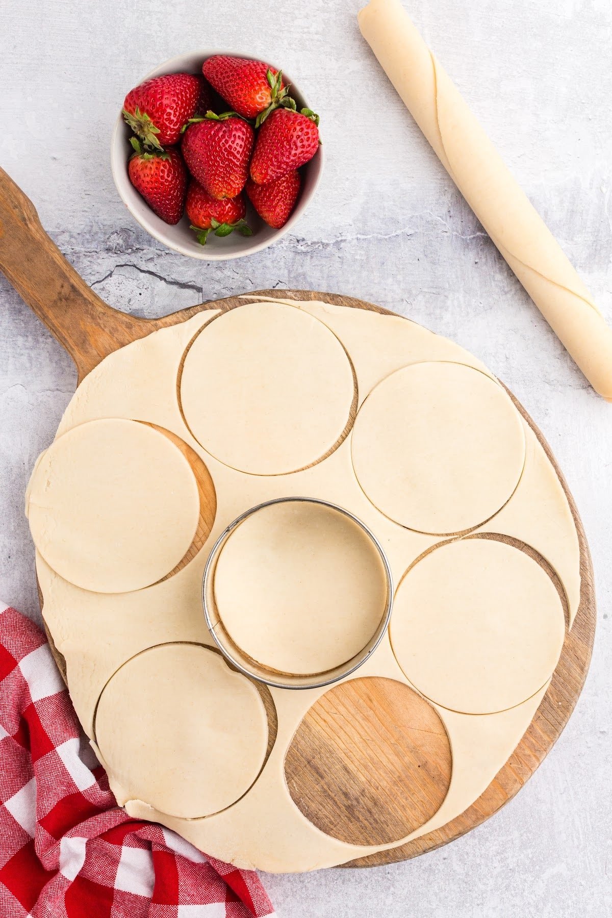 Cutting the pie dough using a round cookie cutter, on a cutting board with strawberries int he background.