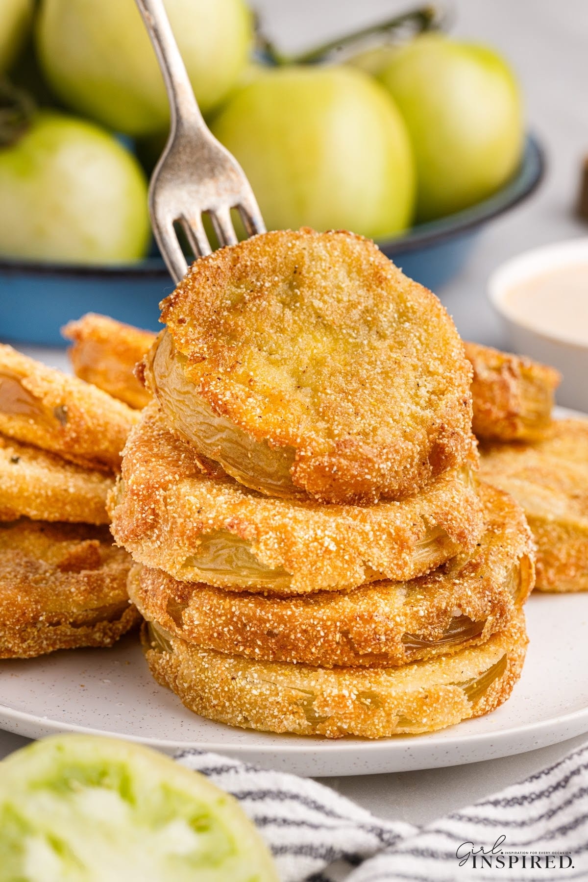 Stack of fried green tomatoes on a platter, being served with a fork, and a bowl of green tomatoes in the background.