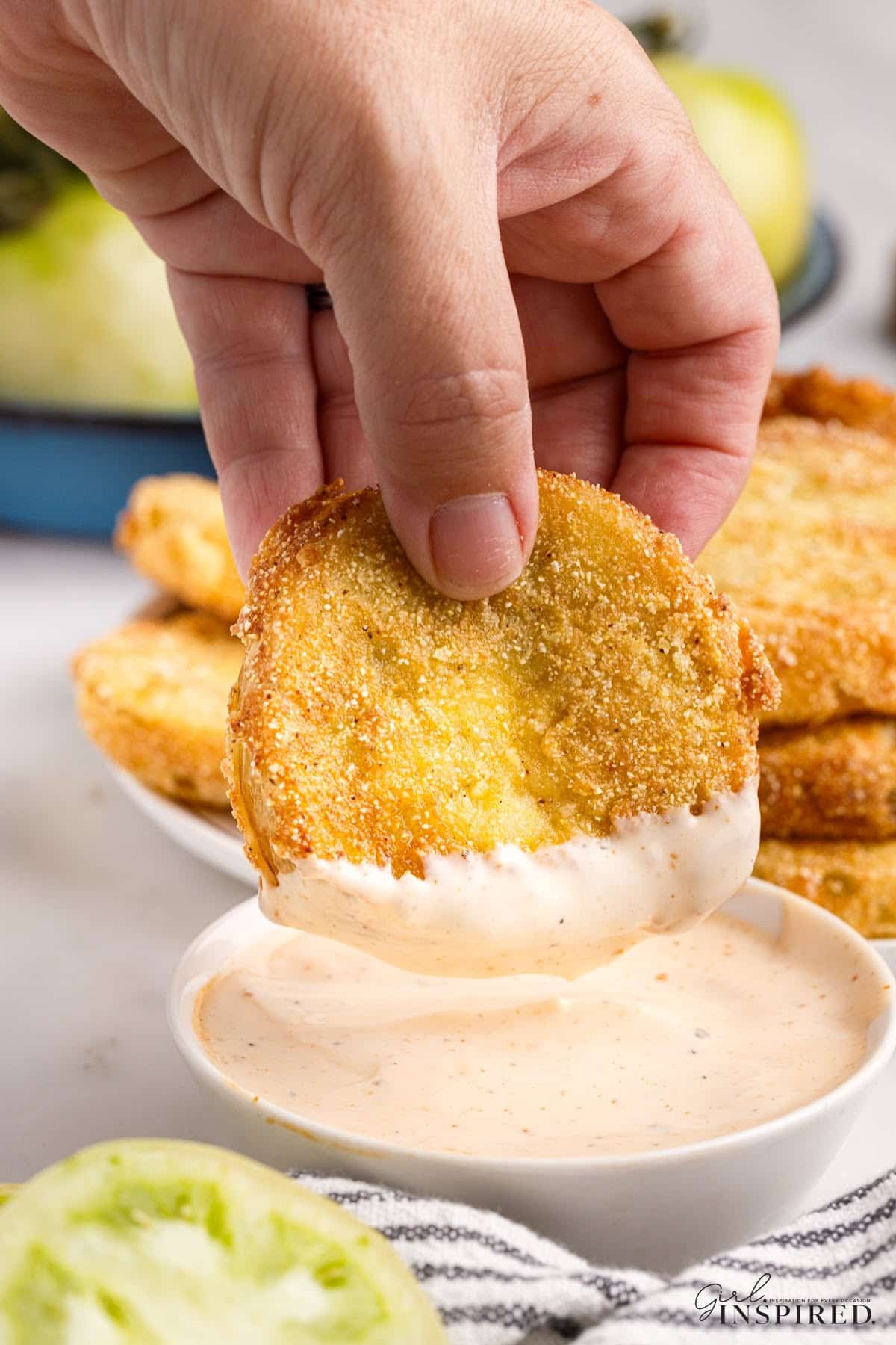 A fried green tomato being dipped in sauce, with a stack of other fried green tomatoes in the background.