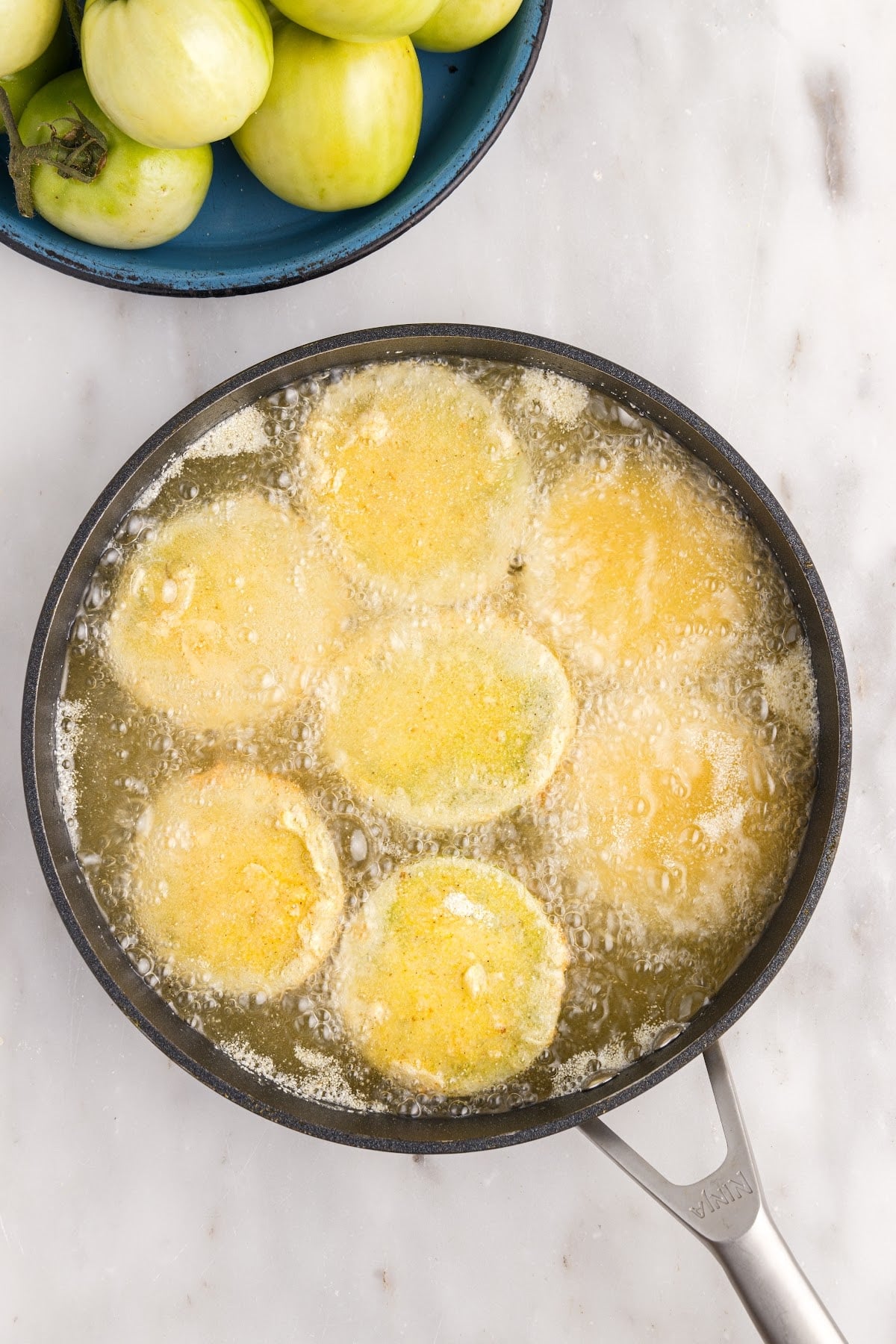 In a single layer, frying the tomatoes in the oil, a bowl of green tomatoes off to the side.
