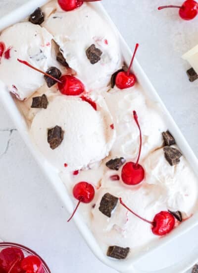 Cherry Garcia ice cream mounded in a loaf pan with maraschino cherries and chocolate chunks scattered over the top and on the table beside some ice cream cones.