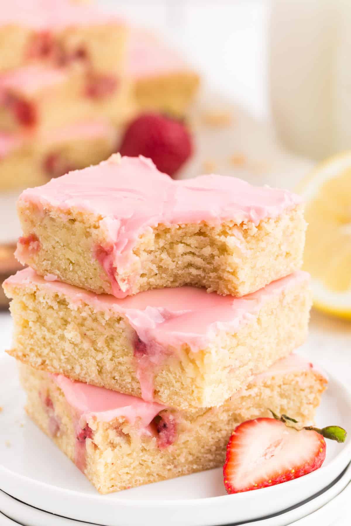 Tall stack of Strawberry Lemon Blondies on a plate showing the berries and brownie inside with pink frosting and a bite taken out of the top one.