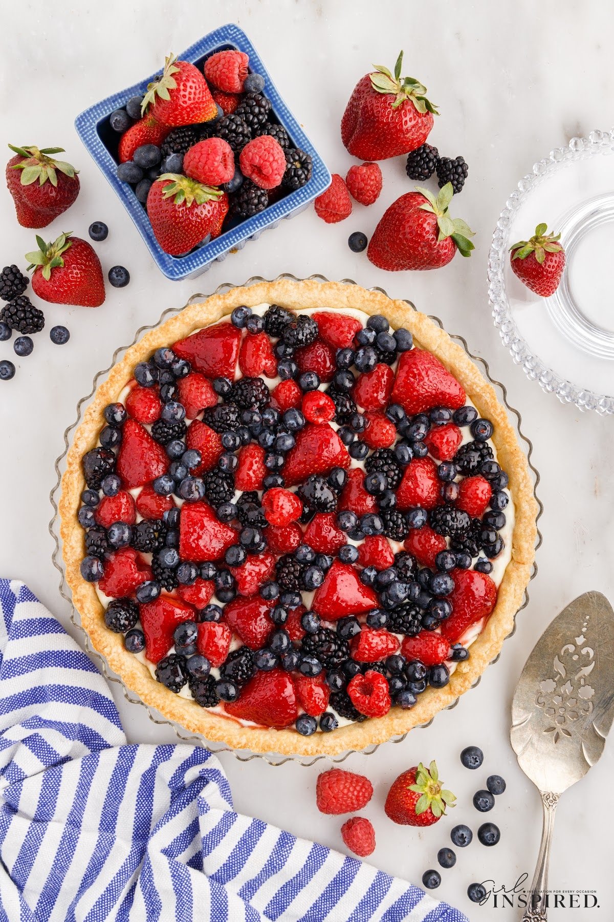 Mixed berry tart with additional fresh berries scattered around, a blue-striped kitchen cloth to the side of the tart with a silver server.