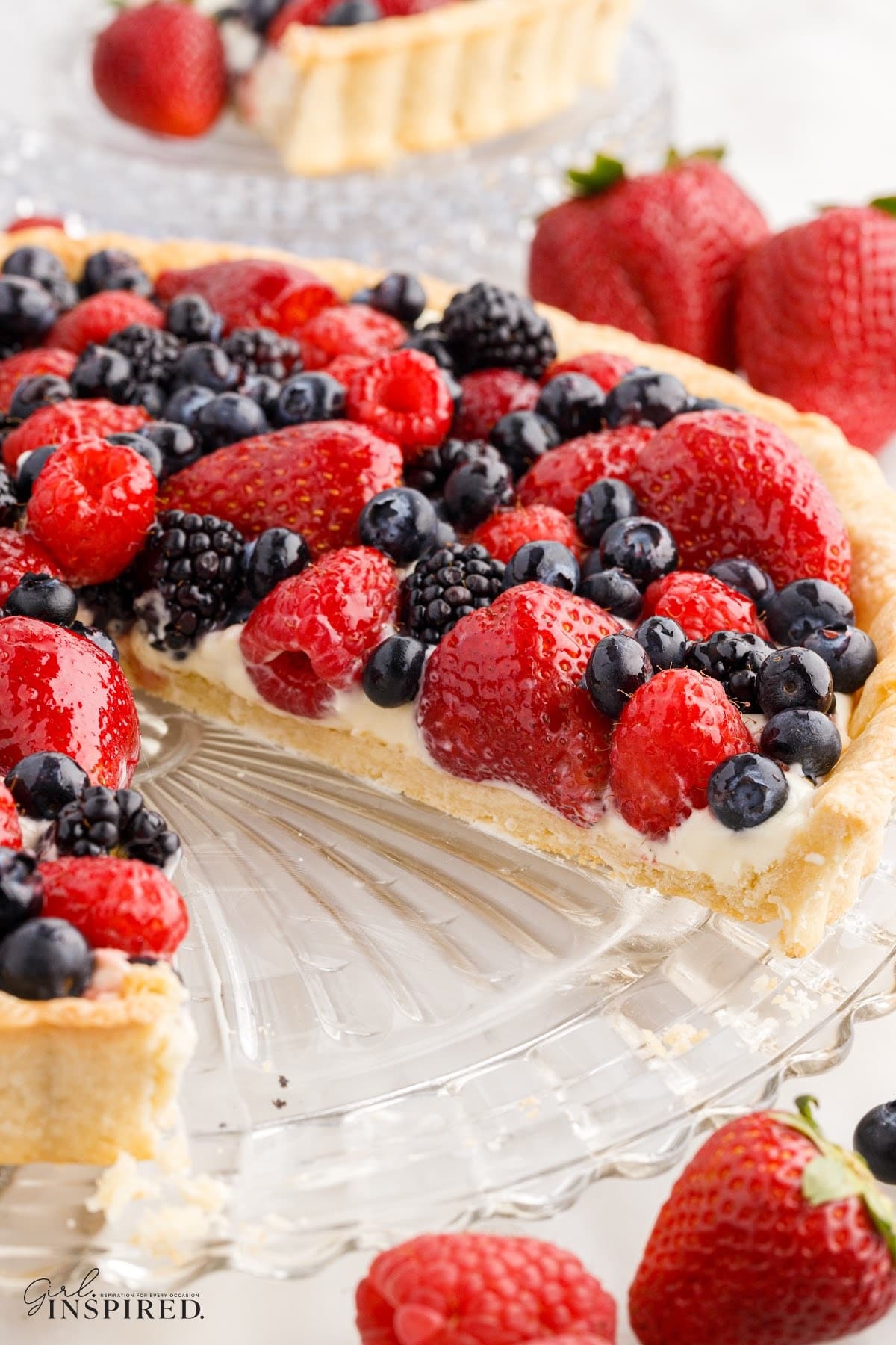 Mixed Berry Tart with slice of tart removed, fresh berries scattered around, a single slice of the tart on a serving plate in the background.