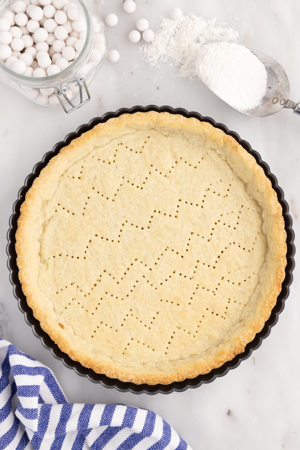 Overhead view of the baked pastry shell, blue and white striped kitchen cloth, jar of pie beads, and sugar scoop.