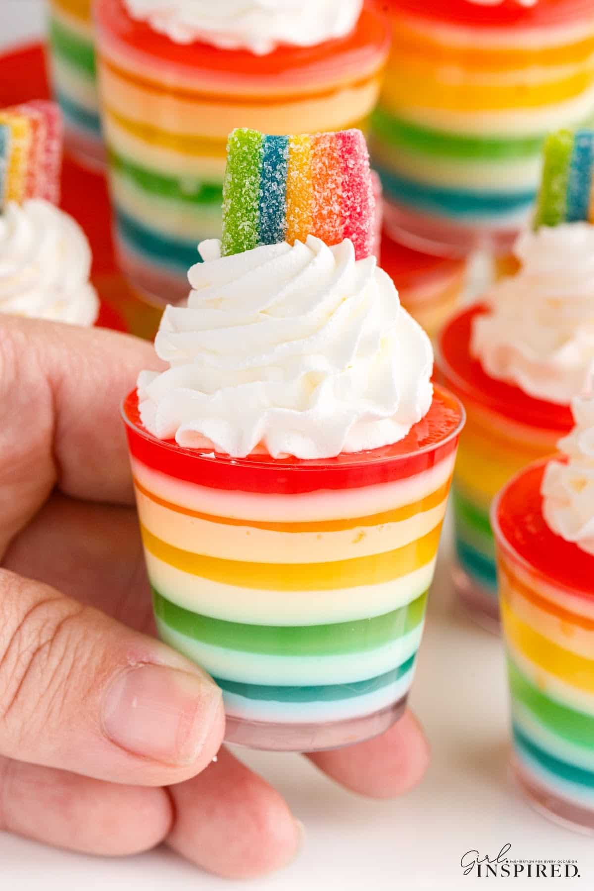 Hand holding a rainbow jello shot, additional jello shots in the background.