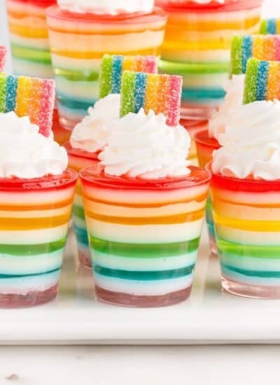 Rainbow Jello Shots garnished with whipped cream and rainbow candies on a white serving board.