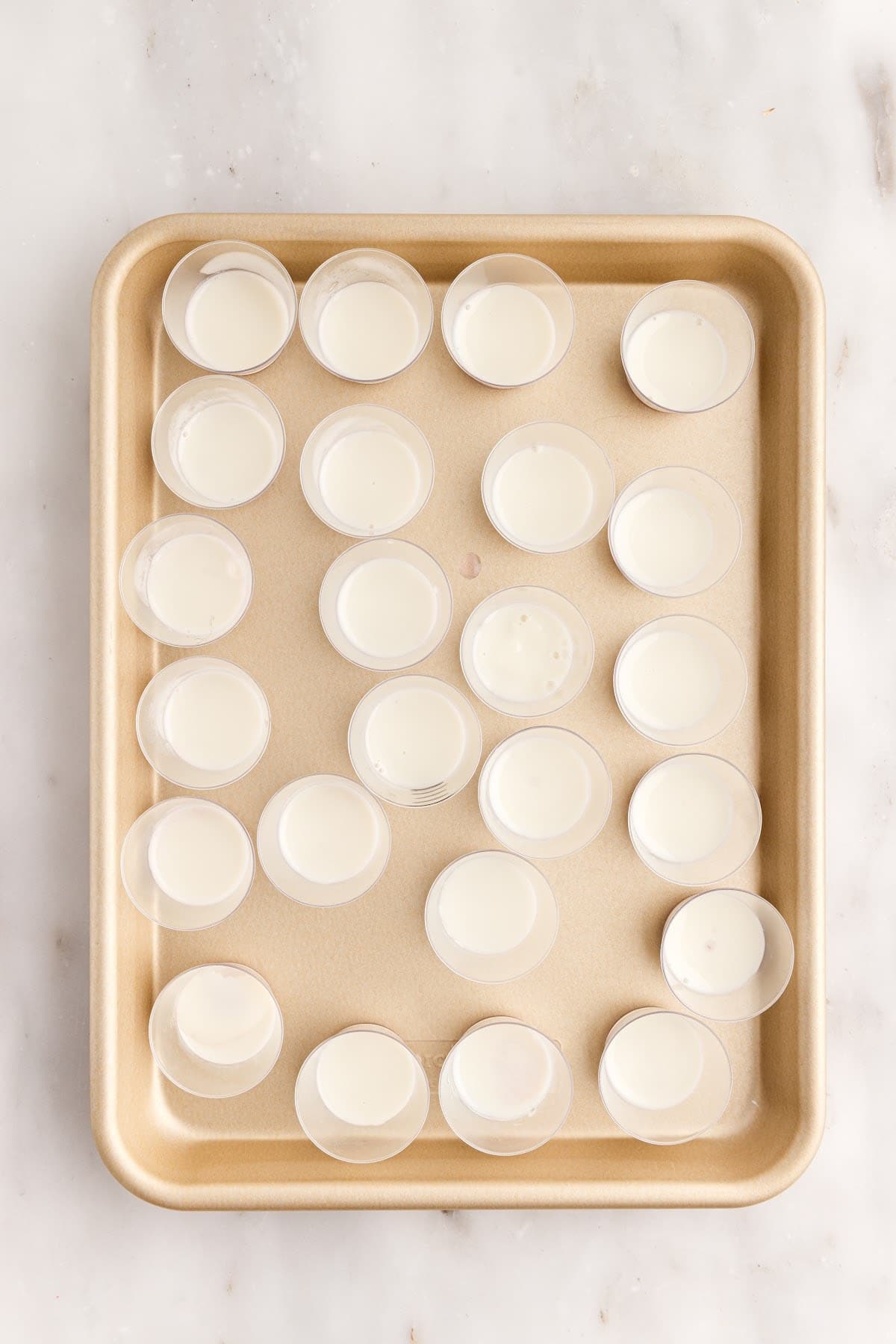A thin layer of the white Jello mixture added to the chilled purple Jello layer in plastic shot glasses on a tray.