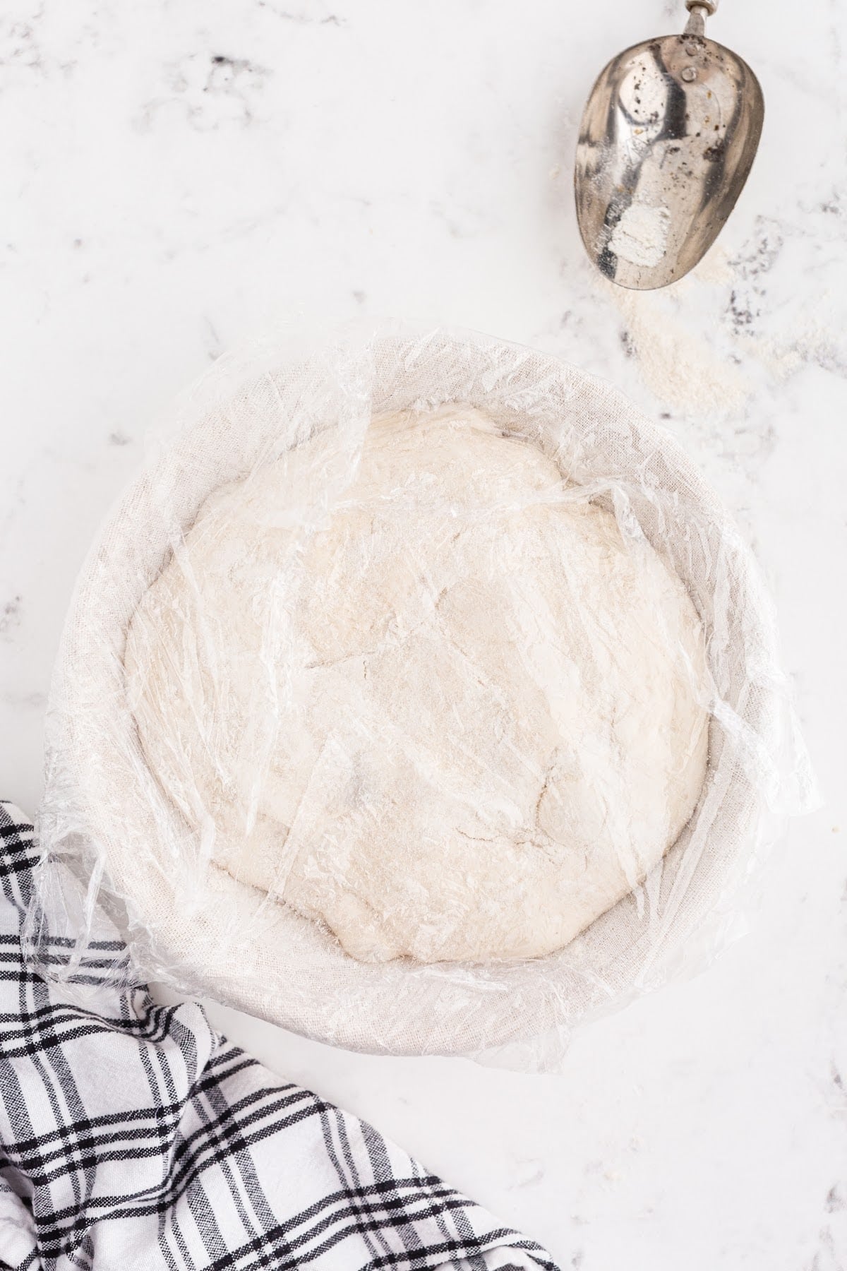 Banneton basket with bread dough wrapped in plastic wrap.