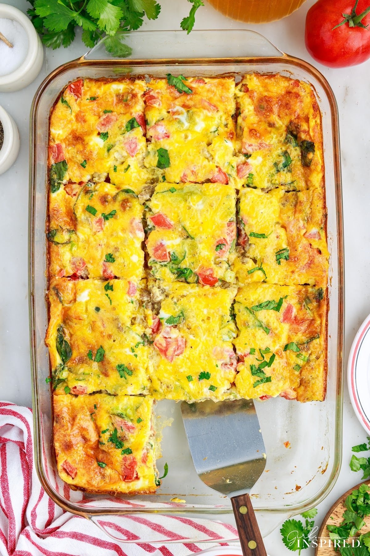 Sliced cheesy egg bake with two servings removed and a silver server. Red striped kitchen cloth and fresh ingredients surrounding the baking dish.