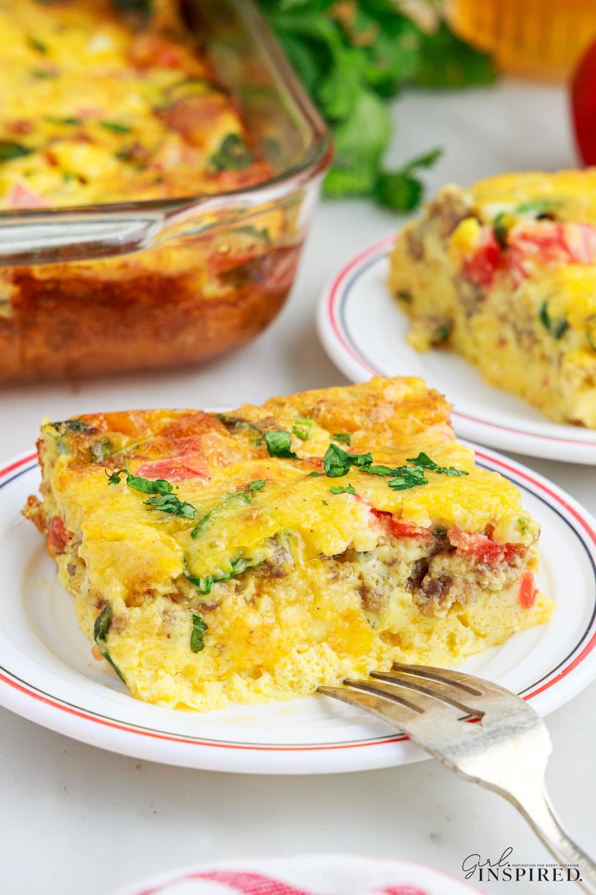 A slice of cheesy egg bake on a white serving plate, additional serving of cheesy egg bake and baking dish in the background.
