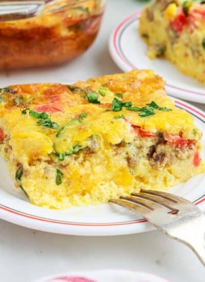 Slice of cheesy egg bake on a white plate with a silver fork, remainder of egg bake in the background.