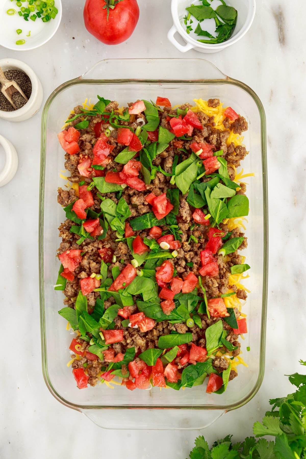 Baking dish with a bottom layer of shredded cheese and a mixture of sausage, chives, tomatoes, and chopped spinach on top, to make the cheesy egg bake.