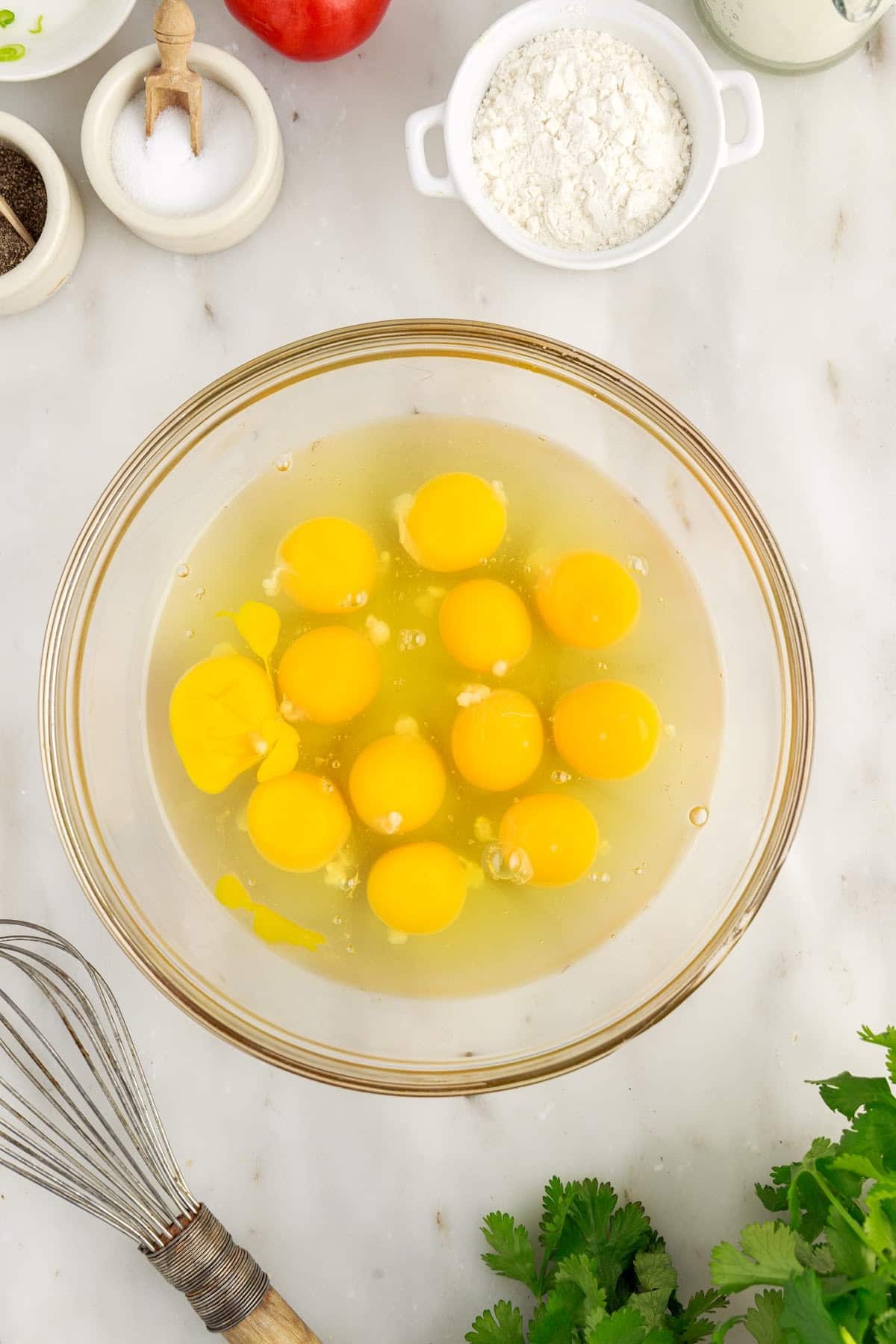 Eggs in a large mixing bowl with metal whisk and remaining ingredients on a marble countertop to make cheesy egg bake.