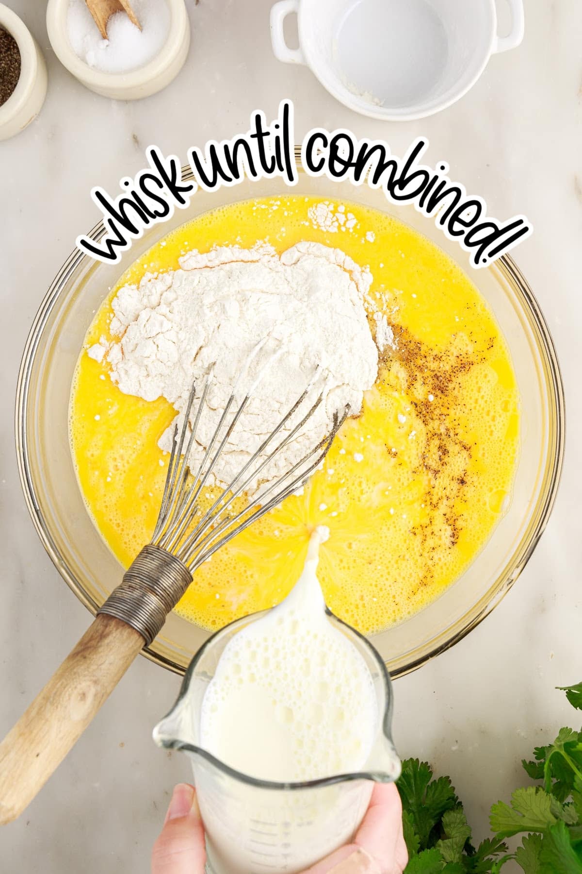 Large mixing bowl with whisked eggs, salt, pepper, and flour with a metal whisk and milk being poured into the bowl.