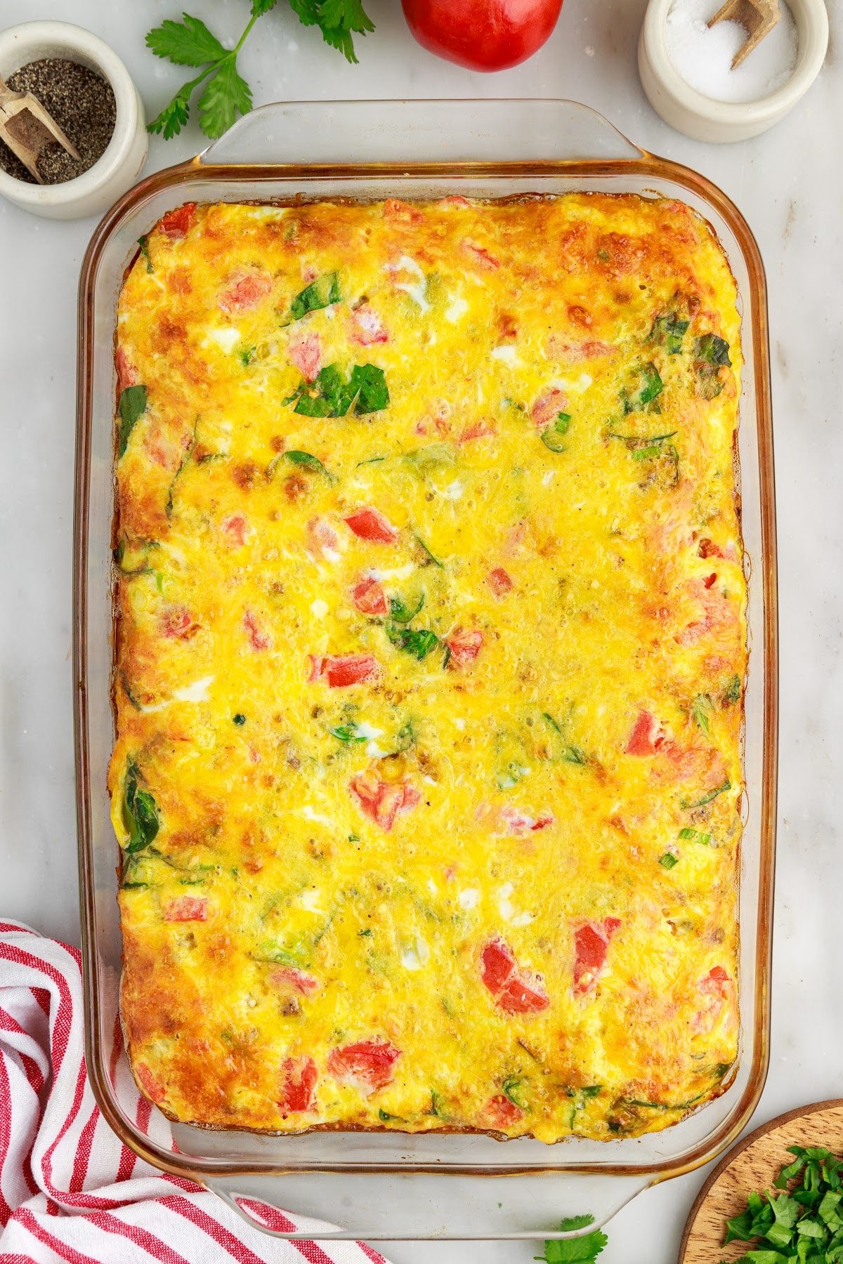 Overhead view of cheesy egg bake with red striped kitchen cloth and leftover ingredients in the background.