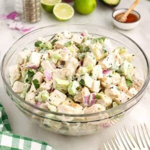 Chicken salad with Greek yogurt in a glass mixing bowl with fresh salad ingredients, forks, and a checkered cloth in the background.