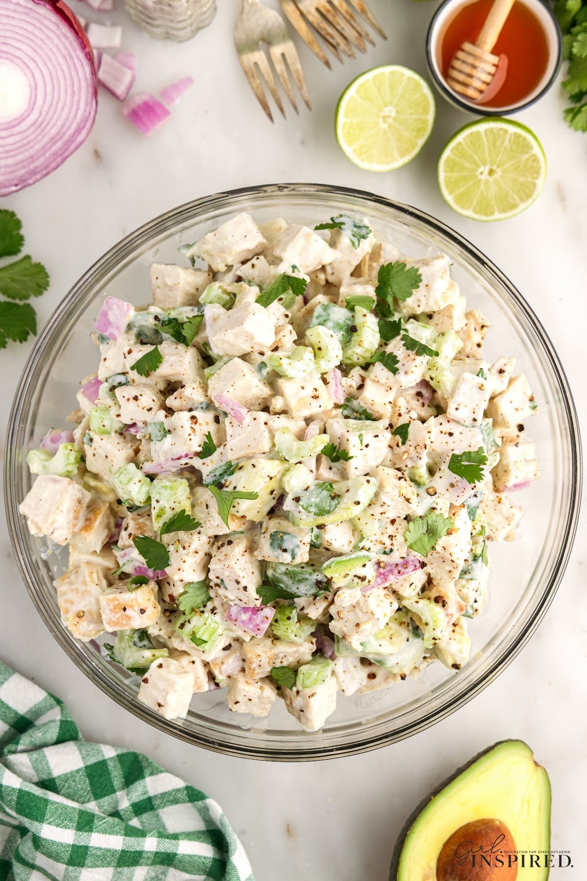 Chicken salad with Greek yogurt dressing in a glass mixing bowl, fresh salad ingredients scattered around with a white and green checkered cloth. 