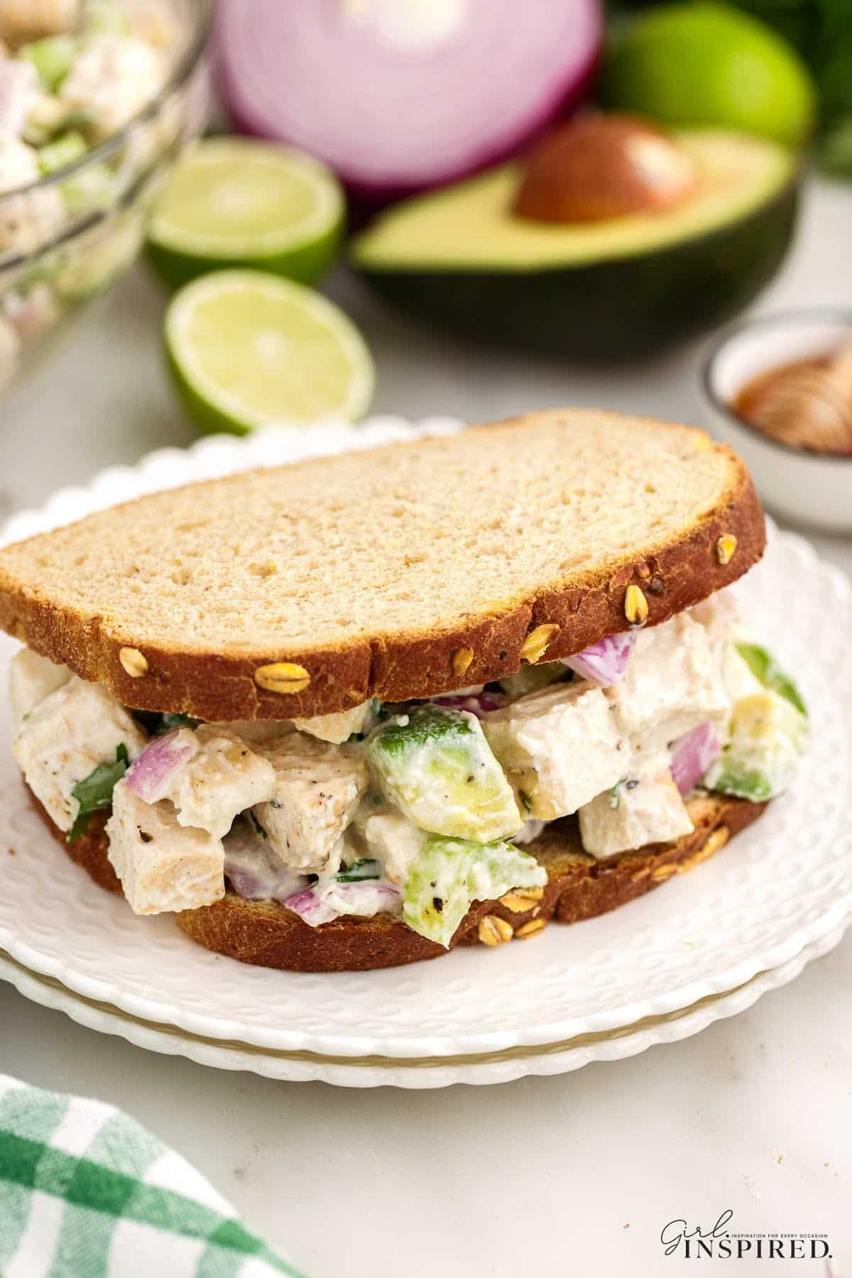 Chicken salad with Greek yogurt loaded into a sandwich and plated on a small serving plate, fresh salad ingredients in the background.