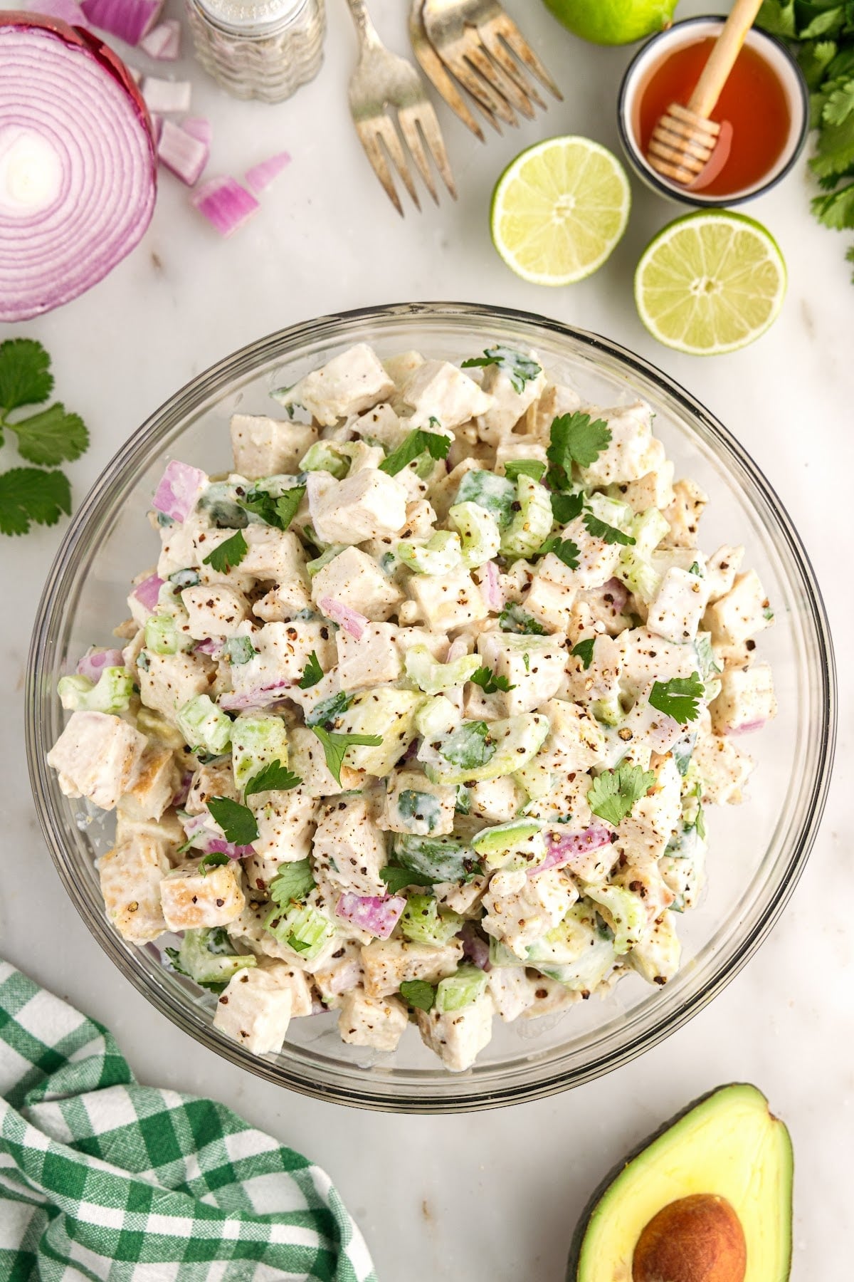 Freshly assembled chicken salad with Greek yogurt dressing in a glass bowl, fresh salad ingredients surrounding the bowl, white and green checkered cloth in the corner.