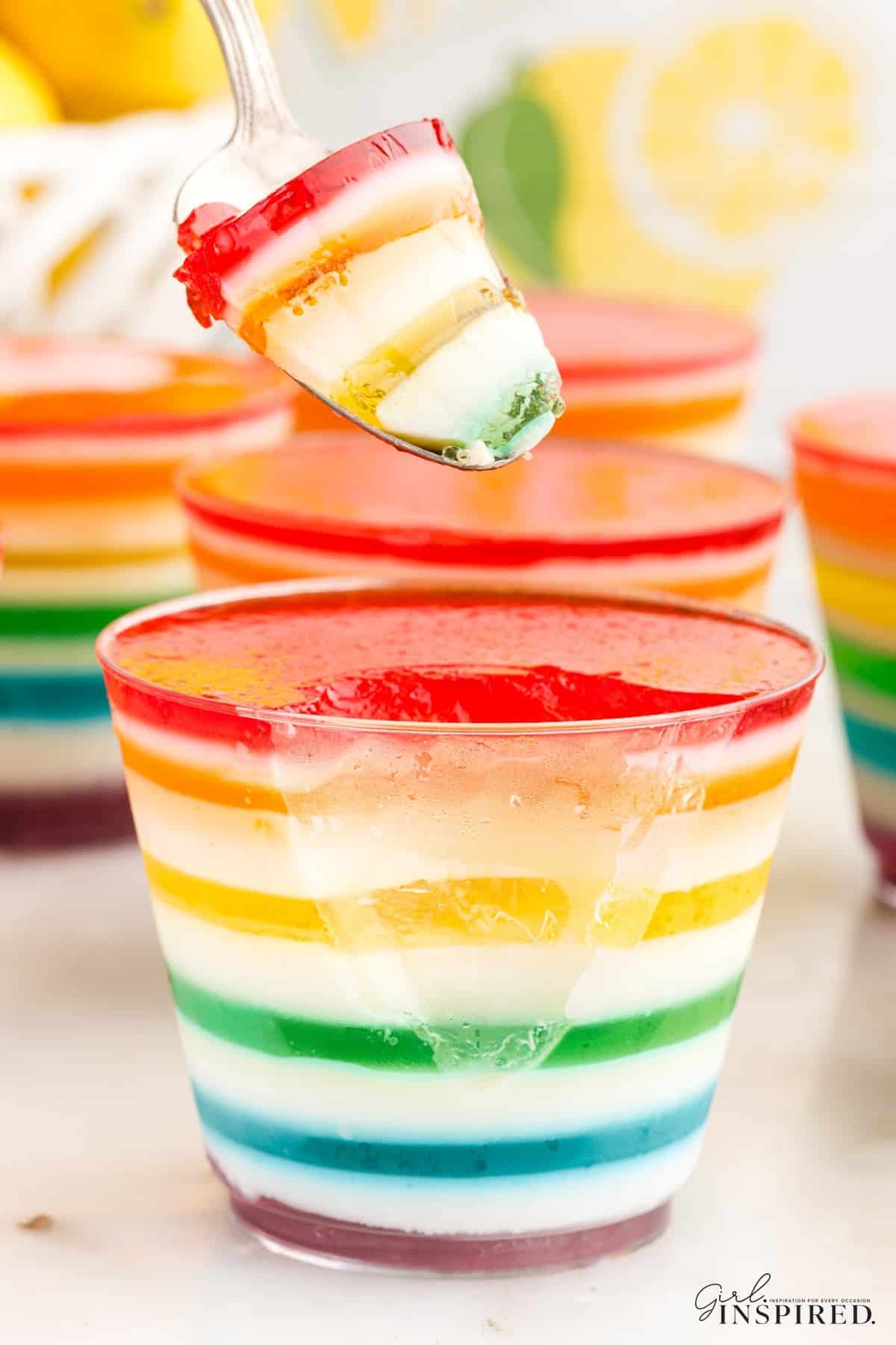 Spoonful of the rainbow layered jello dessert removed from an individual cup. Additional rainbow layered jello cups in the background.