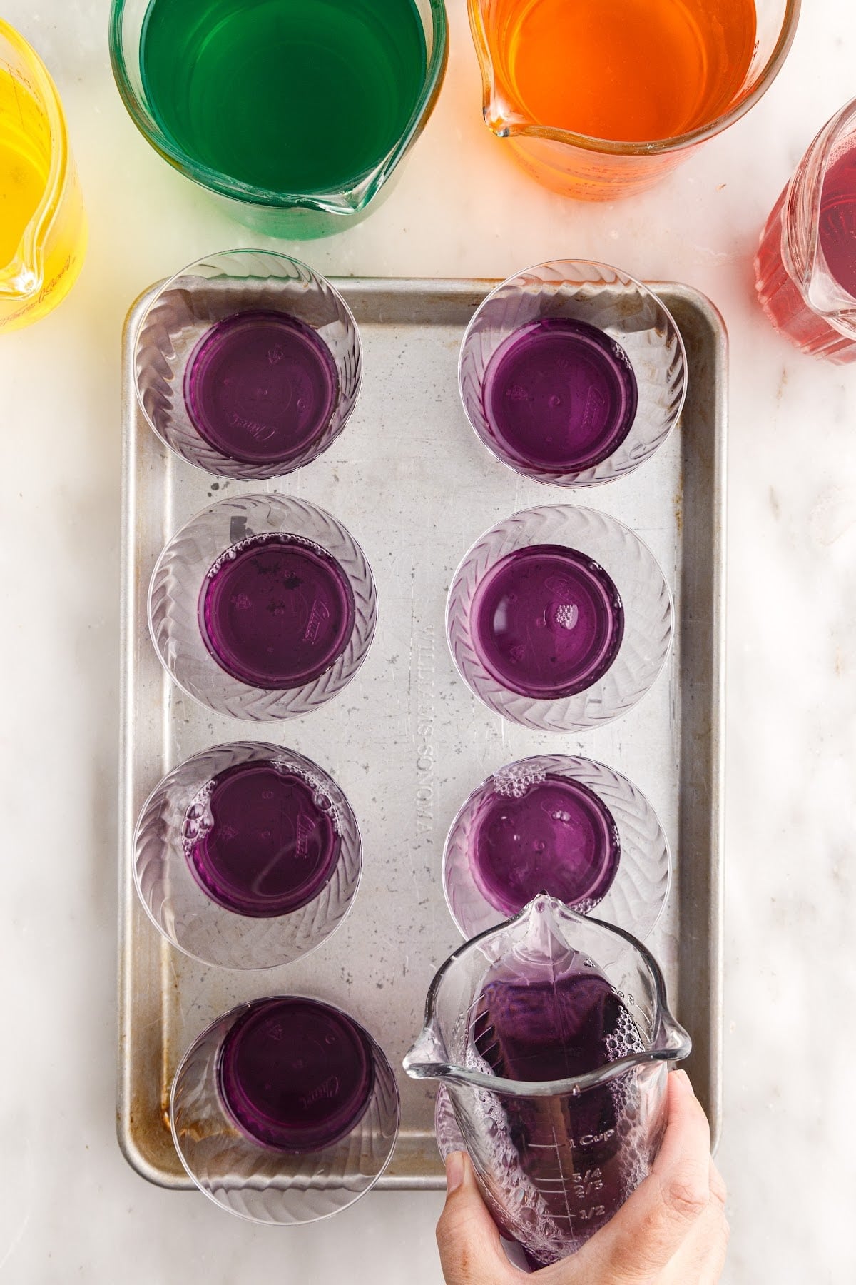 A rimmed baking tray with plastic jello cups and dissolved purple jello added to each cup as the first layer of these rainbow layered jello treats.