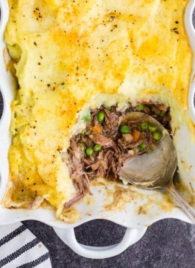 Overhead view of shepherd's pie with leftover roast beef, a single serving removed from the pie dish with a silver serving spoon, striped kitchen cloth alongside the pie dish on a dark marble countertop.