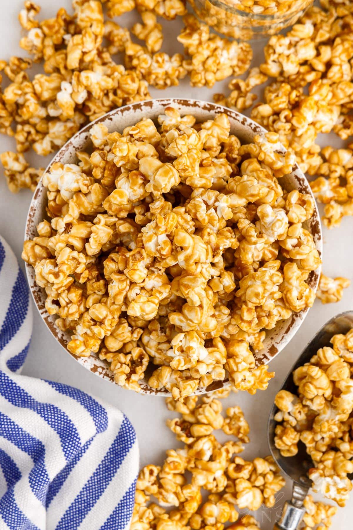 Overehead shot of bowl filled with caramel corn and pieces of caramel corn and blue and white dishtowel sprinkled alongside bowl.