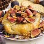 Close up of baked brie with fig jam, topped with quartered figs, chopped candied walnuts, and fresh herbs. Crackers and other fresh ingredients scattered around in the background.