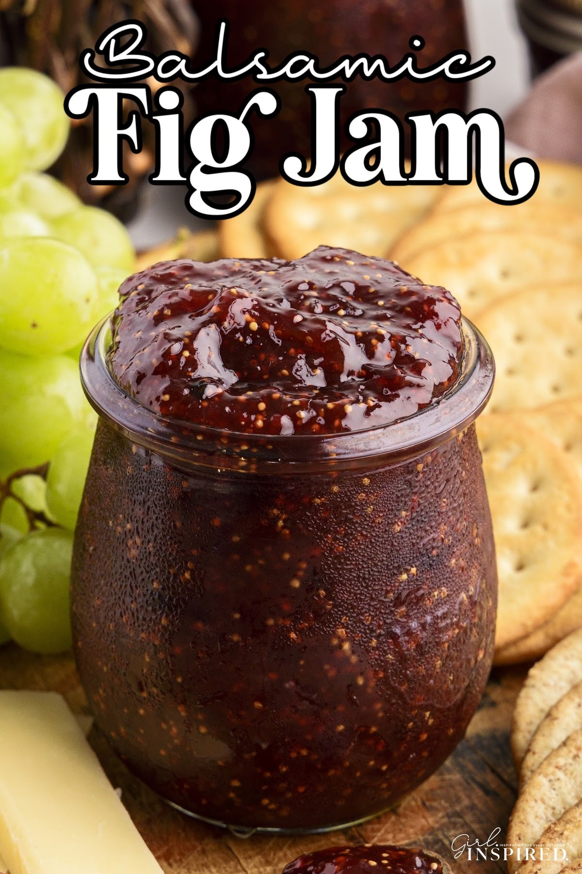 Glass jar filled with homemade balsamic fig jam with crackers, cheese, and fresh fruit placed around.