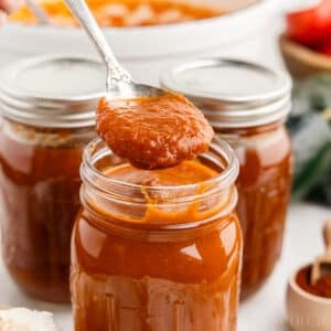 Metal spoon with red enchilada sauce above an open jar of red enchilada sauce, additional jars of sauce in the background.