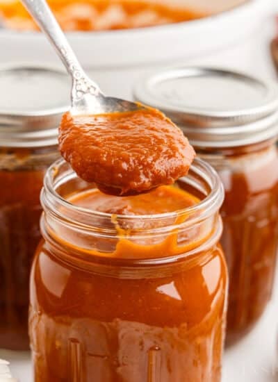 Metal spoon with red enchilada sauce above an open jar of red enchilada sauce, additional jars of sauce in the background.