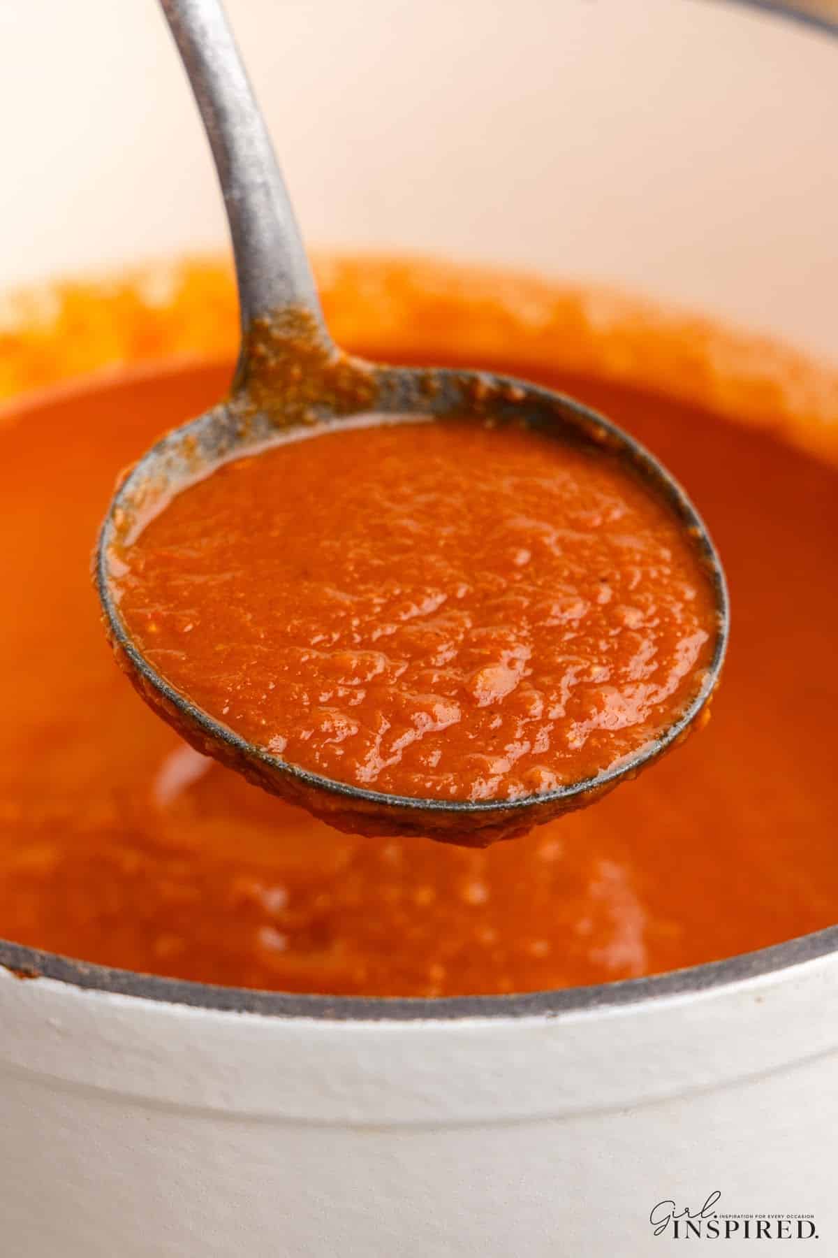 Close up of a metal ladle with red enchilada sauce over a pot of freshly cooked red enchilada sauce.