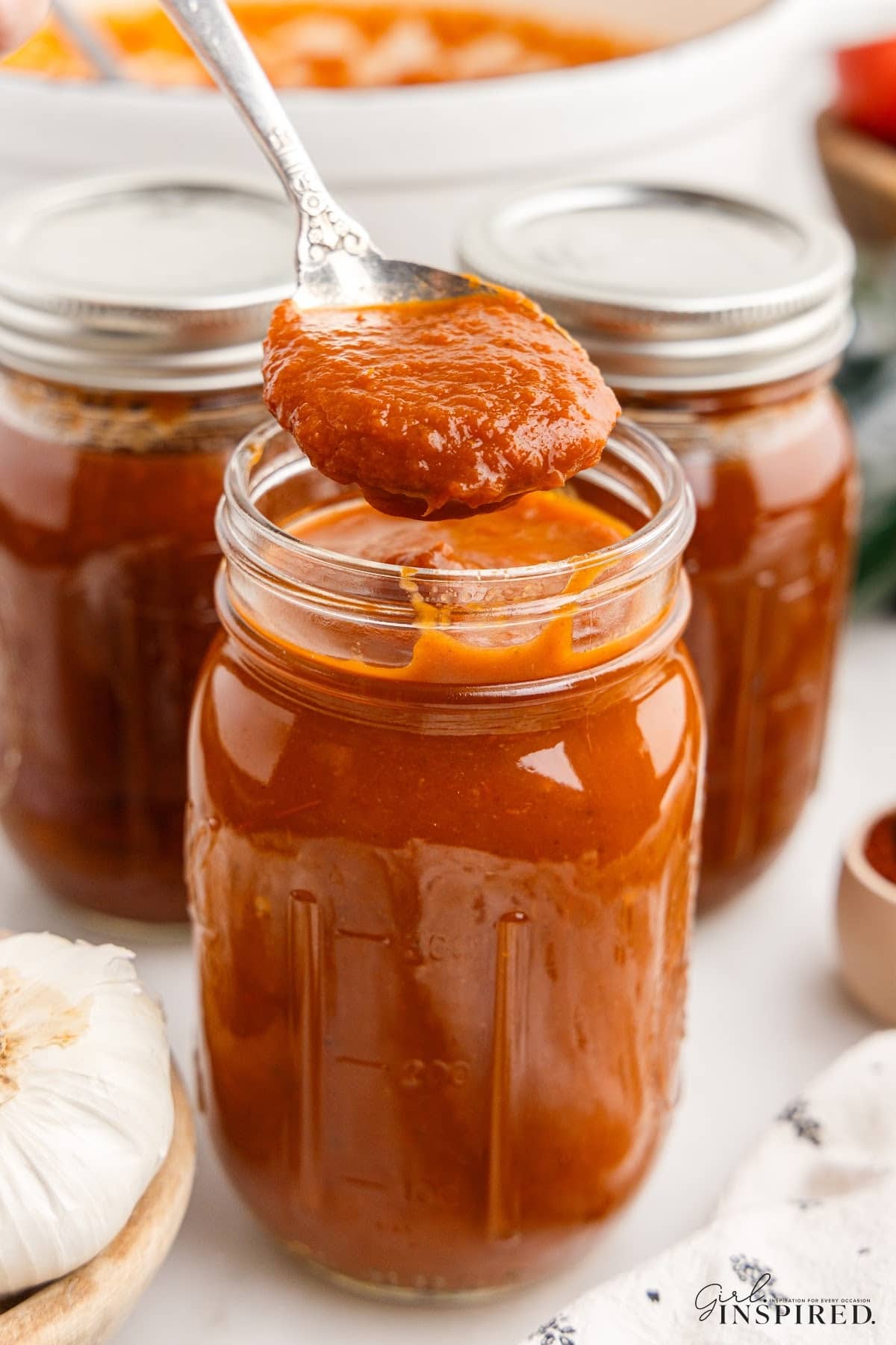 Metal spoon with red enchilada sauce above an open jar of red enchilada sauce, additional jars of sauce in the background.