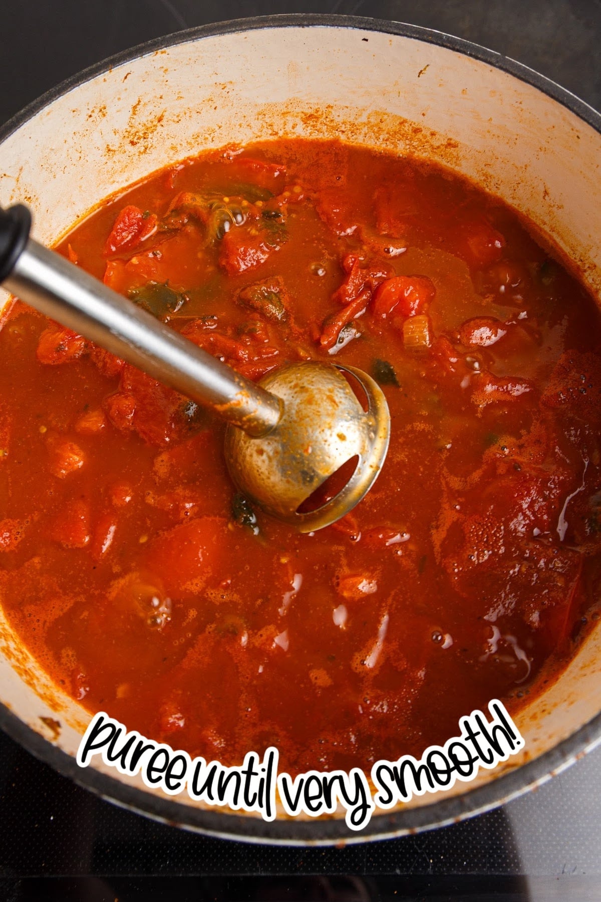 Overhead view of the broken down tomato mixture with an immersion blender, blending the red enchilada sauce mixture until smooth.